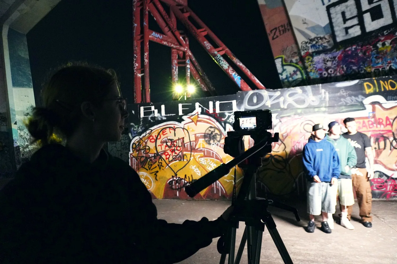 Cámara en trípode frente a una pared de grafiti, con tres personas posando en el rodaje nocturno