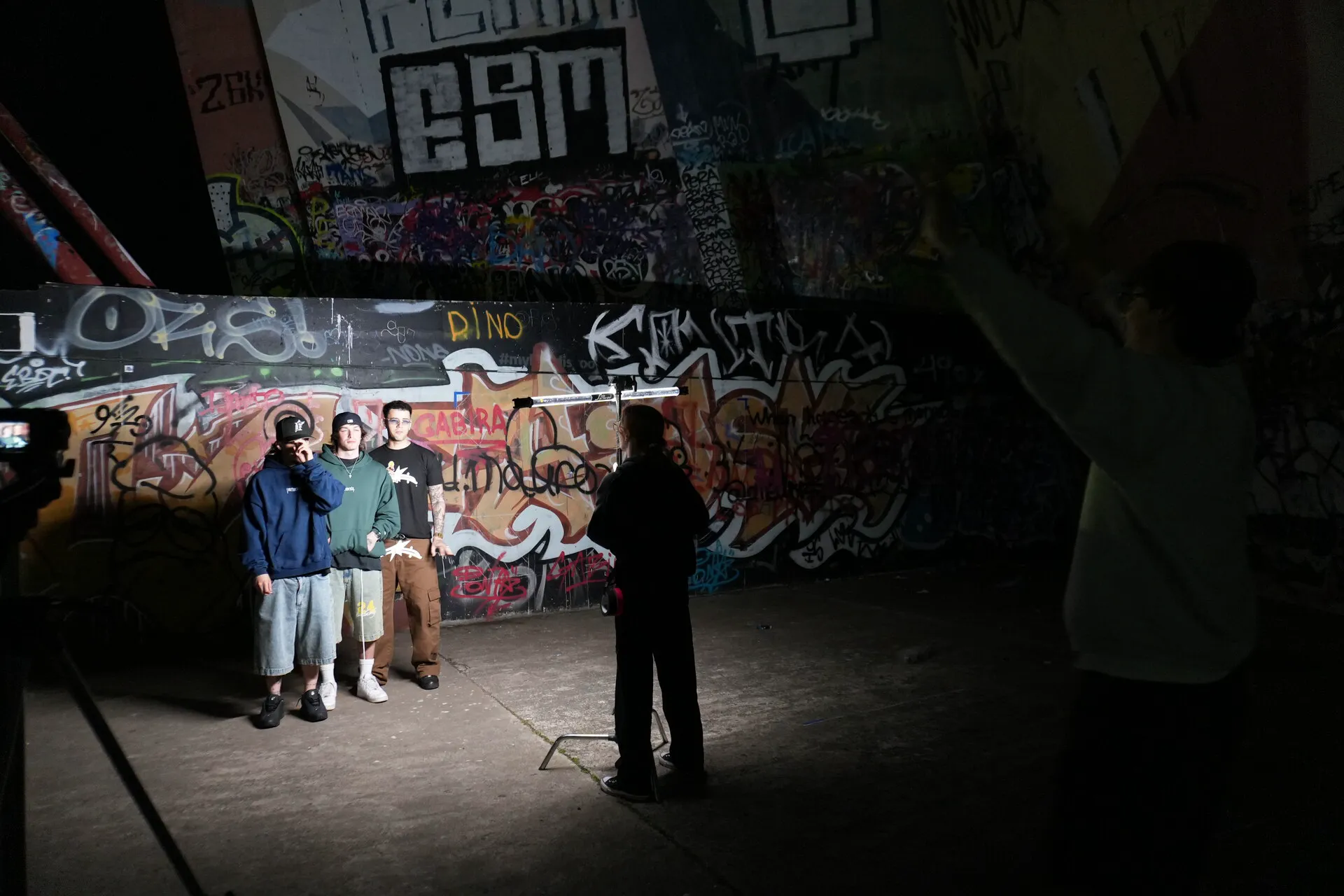 Rodaje nocturno de Kickout: tres artistas posan frente a una pared de grafiti bajo un foco, con parte del equipo en primer plano